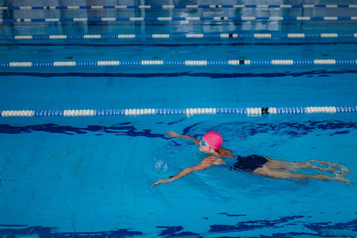 eldre dame med rosa badehette og svømmebrille trener i et offentlig basseng. - Klikk for stort bilde