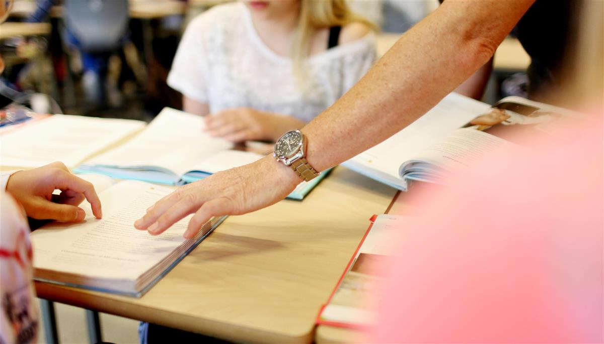 utsnitt av et klasserom som viser pullt med bok og en hånd oppå boka. Det er også en hånd som peker på boka. - Klikk for stort bilde